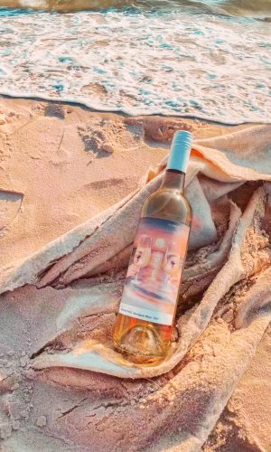 bottle of white wine with hand painted label lays on the white sand of the beach. Waves nearly touching the bottle. Golden rays of sunlight shines on the wine, sand and sea.