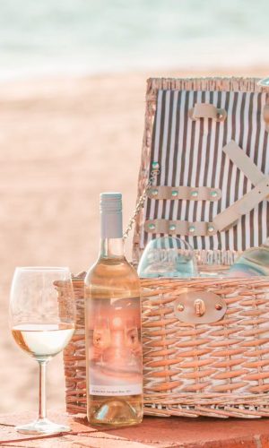 straw picnic basket sits on the sand of the beach. Waves rolling in the distance. Bottle and glass of white wine sit in front of picnic basket