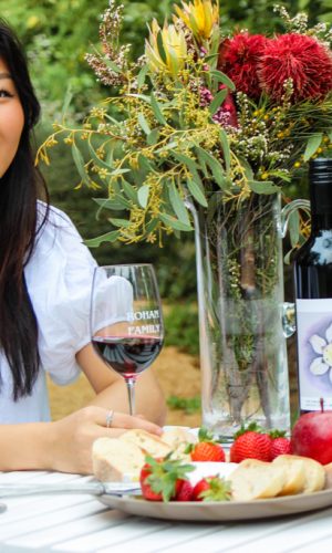 Girl in white dress sits at white wooden table in a luscious green garden. She has in her hand a glass of Shiraz.