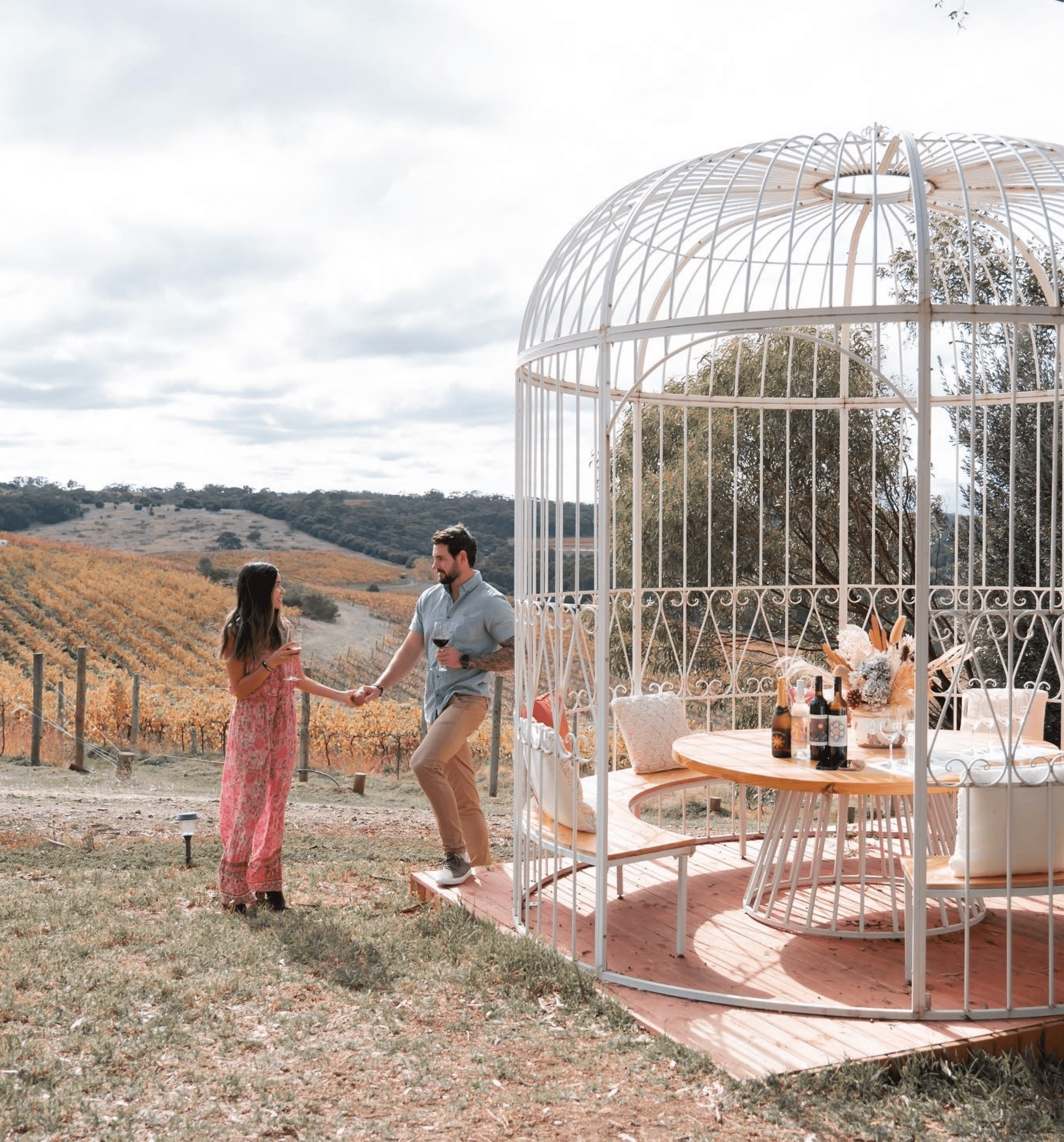 man & woman drinking wine in front of birdcage seating area at winery with vineyard in the background